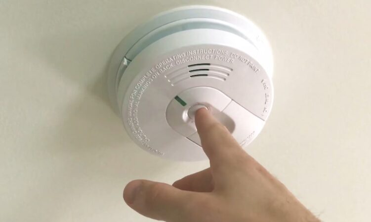 A person reaching up to touch a smoke detector mounted on a wall