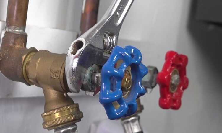 Close-up of a chrome adjustable wrench tightening a brass fitting on a plumbing valve with a bright blue handle, next to a red handle