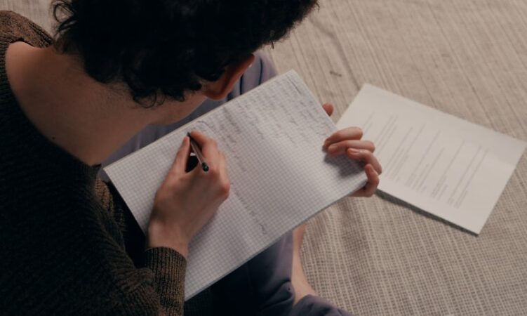 A person sitting on the floor, focused on writing in a notebook with a pen